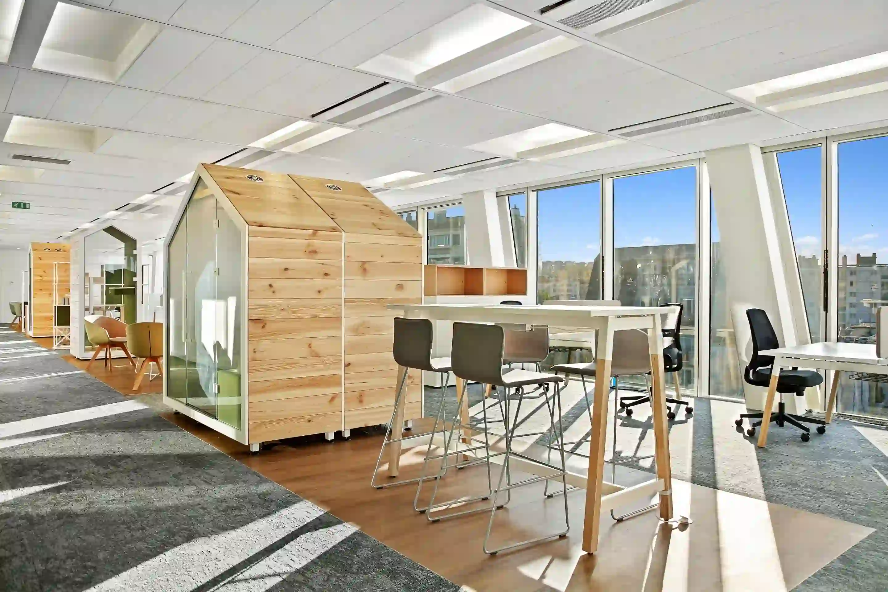 Espace de bureau lumineux et ouvert avec cabines individuelles en bois en forme de maisonnette, table haute avec tabourets, et grandes baies vitrées donnant sur la ville